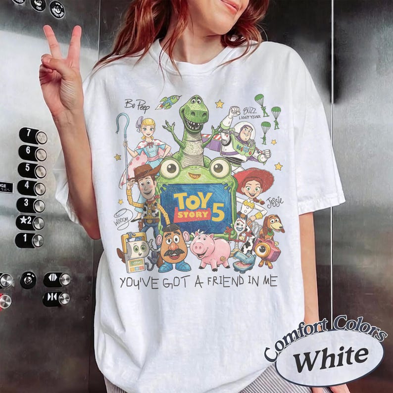 May include: A white short-sleeve t-shirt featuring a colorful graphic print of various animated characters from Toy Story. The design includes the text "TOY STORY 5" and "YOU'VE GOT A FRIEND IN ME" along with illustrations of Woody, Buzz Lightyear, Jessie, Rex, and other characters.