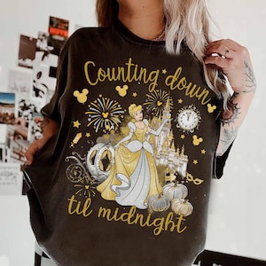 Può includere: T-shirt grigio scuro con un design a tema Cenerentola e la scritta "Counting down til midnight". La grafica include Cenerentola, una carrozza a zucca, fuochi d'artificio, un castello e zucche in oro e bianco.