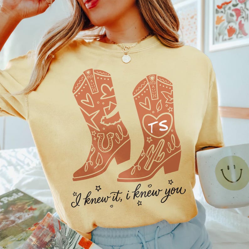 May include: A pale yellow t-shirt featuring a graphic of two orange cowboy boots decorated with hearts, stars, and a horseshoe. One boot displays the letters TS inside a heart. The text "I knew it, I knew you" is printed in black below the boots.