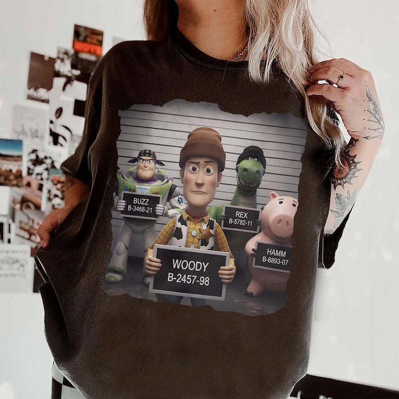 Puede incluir: Una camiseta negra con un gr&aacute;fico de los personajes de Toy Story Buzz, Woody, Rex y Hamm en una fila de identificaci&oacute;n policial. Cada personaje sostiene un cartel con su nombre y un n&uacute;mero de identificaci&oacute;n &uacute;nico sobre un fondo de l&iacute;neas.