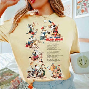 Puede incluir: Camiseta amarilla clara con un dise&ntilde;o vintage del concierto de la banda Mickey Mouse de Walt Disney. El gr&aacute;fico incluye a Mickey Mouse, el Pato Donald y Goofy tocando instrumentos. El texto de la camiseta dice "Mickey Mouse The Band Concert".
