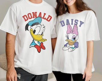 May include: White t-shirts with Donald and Daisy Duck designs. Donald's shirt has his name in red above his image, wearing a blue sailor hat and red bow tie. Daisy's shirt has her name in purple, with a pink bow and purple dress.
