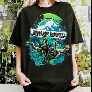 May include: Black t-shirt featuring the "Jurassic World" logo with a dinosaur silhouette and the text "JURASSIC WORLD". Below the logo are illustrations of several dinosaurs in a jungle setting.
