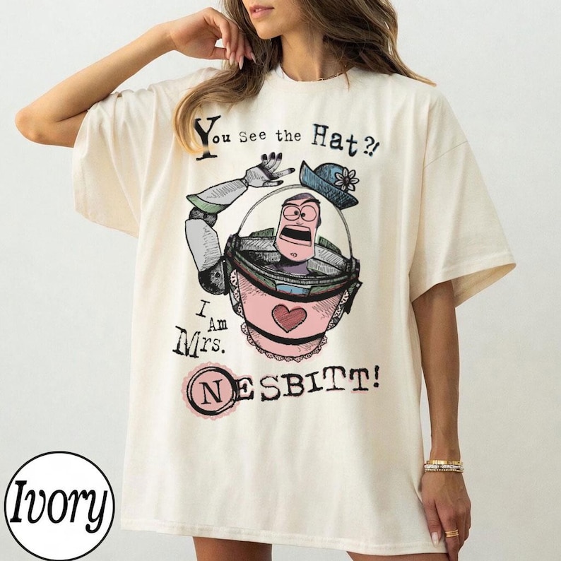 Puede incluir: Camiseta de color marfil con una ilustraci&oacute;n de dibujos animados de un personaje con un vestido rosa y un sombrero azul, con el texto "You See the Hat?!" y "I Am Mrs. Nesbitt!" impreso en la parte delantera.