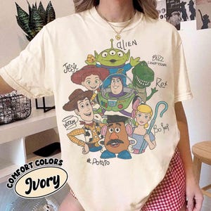 May include: Ivory-colored t-shirt featuring a graphic of characters from the animated film Toy Story. The design includes Woody, Buzz Lightyear, Jessie, Rex, Mr. Potato Head, Bo Peep, and an alien. The shirt also has the words "COMFORT COLORS" and "Ivory".