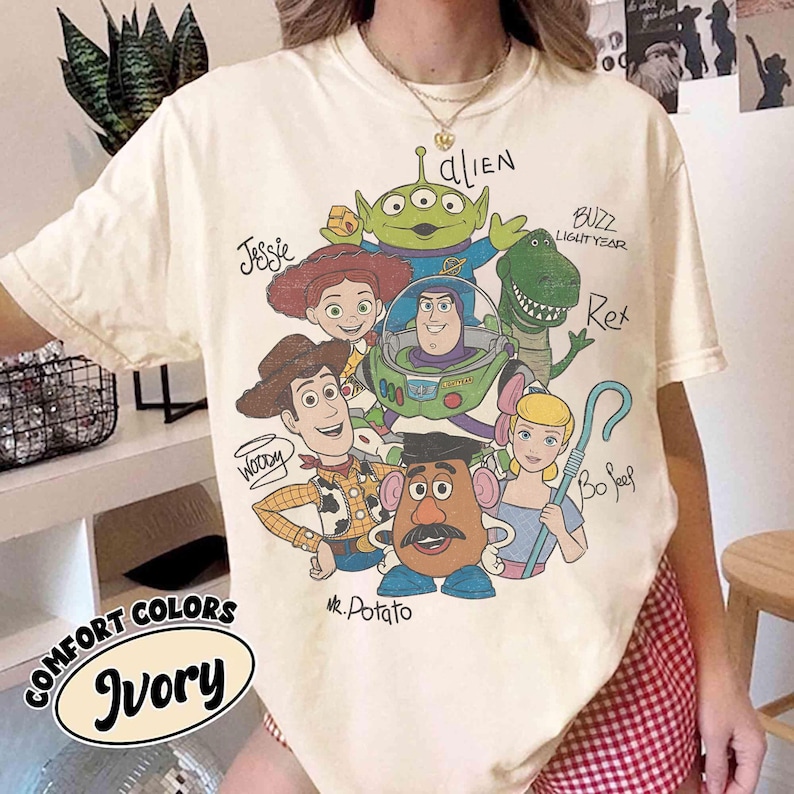 May include: Ivory-colored t-shirt featuring a graphic of characters from the animated film Toy Story. The design includes Woody, Buzz Lightyear, Jessie, Rex, Mr. Potato Head, Bo Peep, and an alien. The shirt also has the words "COMFORT COLORS" and "Ivory".