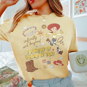 May include: A yellow t-shirt featuring a graphic print of the character Jessie from Toy Story, a sheriff badge, a beaded bracelet with the text 'You've got a friend in me', cowboy boots, blocks with letters, a hat, and the text 'infinity and beyond' and 'I KNEW IT I KNEW YOU'.