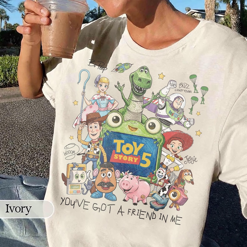 May include: An ivory t-shirt featuring a colorful graphic print of Toy Story characters including Woody, Buzz Lightyear, Jessie, Rex, and Bo Peep. The design includes the text Toy Story 5 and You've Got A Friend In Me, along with various small icons.