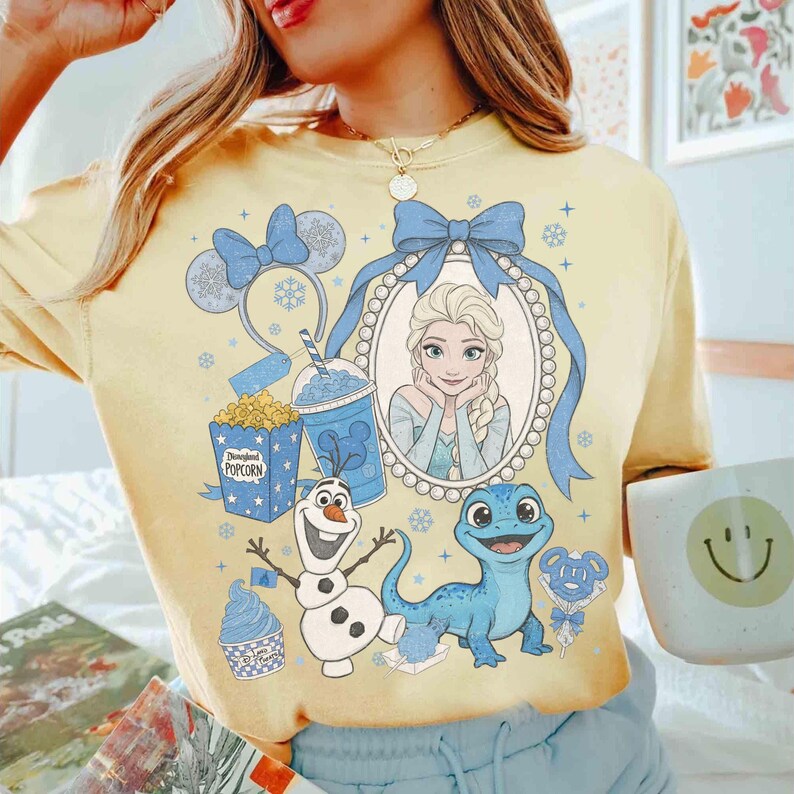 Op de afbeelding: Geel T-shirt met een ontwerp met Elsa uit Frozen, blauwe accenten en Disney-thema graphics. Inclusief Olaf, een blauwe hagedis, popcorn en een drankje. Het shirt heeft een relaxte pasvorm.