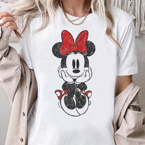 May include: White t-shirt with a vintage-style graphic of Minnie Mouse wearing a red bow and a black dress.