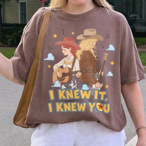 May include: A brown short-sleeve t-shirt featuring a graphic of two cowgirls playing acoustic guitars. The design includes stars, clouds, musical notes, and the text "I KNEW IT, I KNEW YOU" with a red star icon replacing the letter O.
