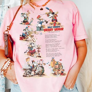 Puede incluir: Camiseta rosa claro con un gr&aacute;fico de dibujos animados vintage de Mickey Mouse y sus amigos tocando instrumentos musicales. El texto "WALT DISNEY'S MICKEY MOUSE The Band Concert" est&aacute; impreso en la camiseta. La camiseta es de material suave.