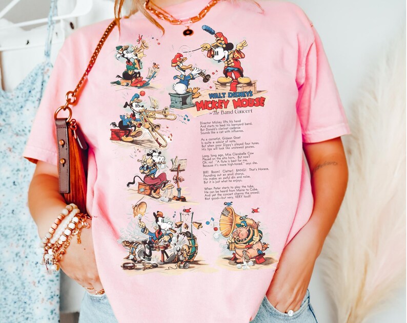 Puede incluir: Camiseta rosa claro con un gr&aacute;fico de dibujos animados vintage de Mickey Mouse y sus amigos tocando instrumentos musicales. El texto "WALT DISNEY'S MICKEY MOUSE The Band Concert" est&aacute; impreso en la camiseta. La camiseta es de material suave.