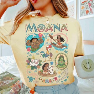 May include: Yellow t-shirt with the word "MOANA" in colourful letters above cartoon illustrations of characters and scenes from the film. The shirt has a crew neck and short sleeves.