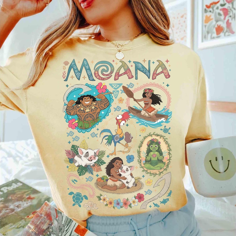 May include: Yellow t-shirt with the word "MOANA" in colourful letters above cartoon illustrations of characters and scenes from the film. The shirt has a crew neck and short sleeves.