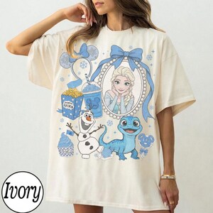 Op de afbeelding: Ivoor kleurig T-shirt met een cartoon ontwerp met Elsa uit Frozen, Olaf en een blauwe hagedis. Het ontwerp bevat een blauwe strik, popcorn en een drankje. Het woord "Ivory" staat onderaan gedrukt.