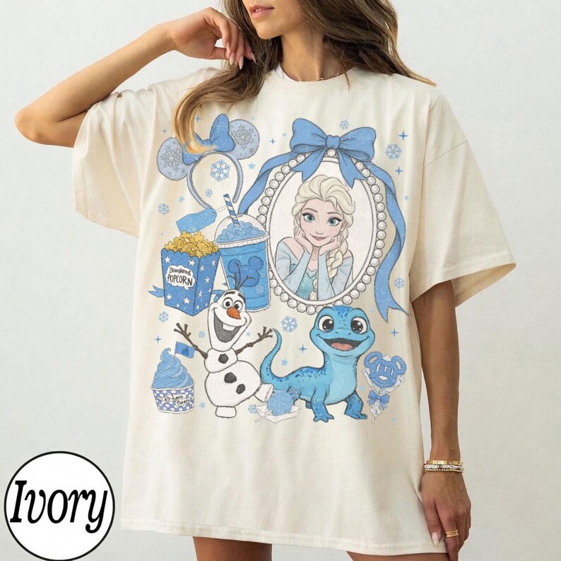 Op de afbeelding: Ivoor kleurig T-shirt met een cartoon ontwerp met Elsa uit Frozen, Olaf en een blauwe hagedis. Het ontwerp bevat een blauwe strik, popcorn en een drankje. Het woord "Ivory" staat onderaan gedrukt.