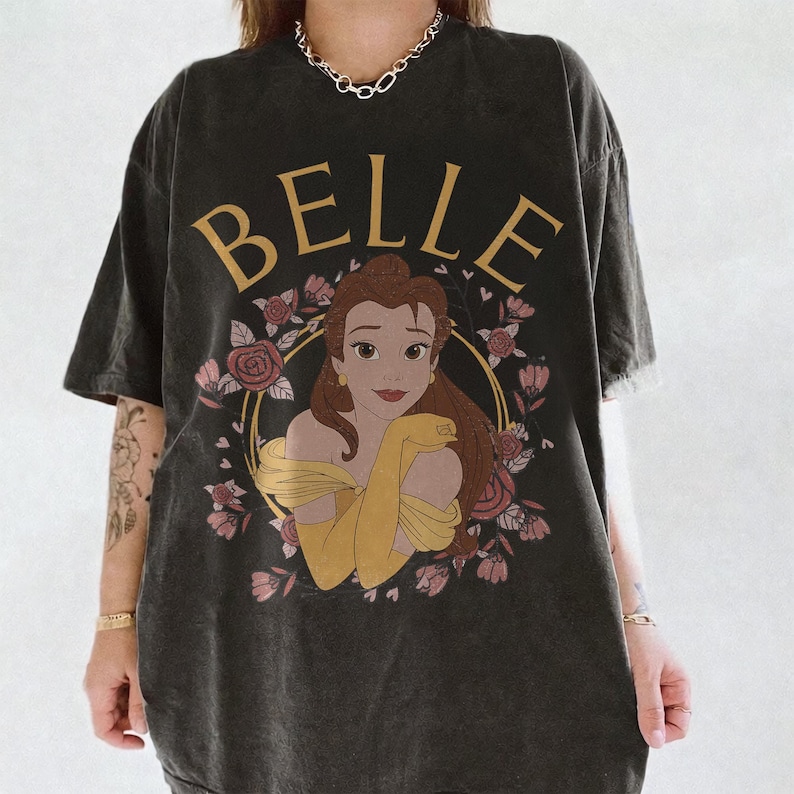 May include: A charcoal gray oversized t-shirt featuring a graphic print of the character Belle from Beauty and the Beast. The design includes the text BELLE in gold lettering above the character, surrounded by a circular frame of pink roses.