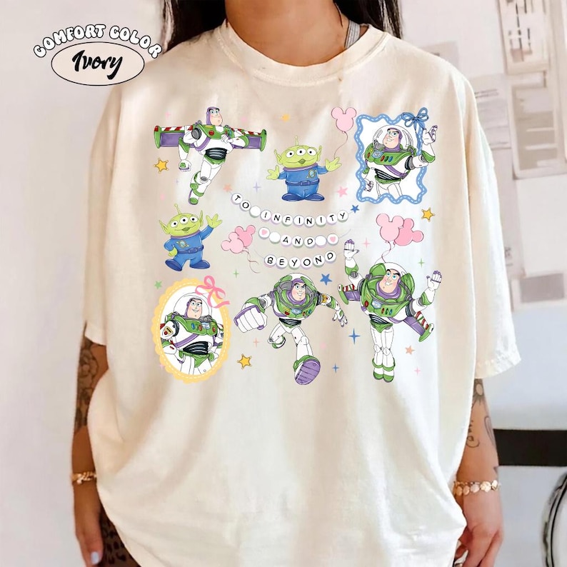 Puede incluir: Camiseta marfil Comfort Color con m&uacute;ltiples im&aacute;genes de Buzz Lightyear y los peque&ntilde;os hombrecillos verdes de Toy Story. La frase "To Infinity and Beyond" tambi&eacute;n est&aacute; presente.