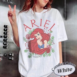 May include: A white short-sleeve t-shirt featuring a graphic of the character Ariel with long red hair, wearing a blue seashell top. The text ARIEL is printed in red above the character, surrounded by green foliage and red coral illustrations.