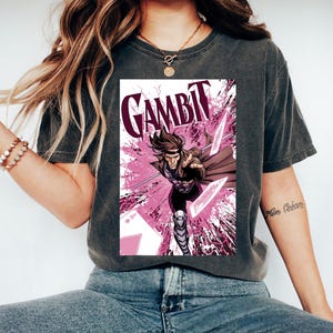 Può includere: T-shirt grigio scuro con grafica di Gambit. L'immagine mostra il personaggio Marvel Comics in azione, su sfondo rosa e bianco. Il nome del personaggio è visualizzato a grandi lettere.