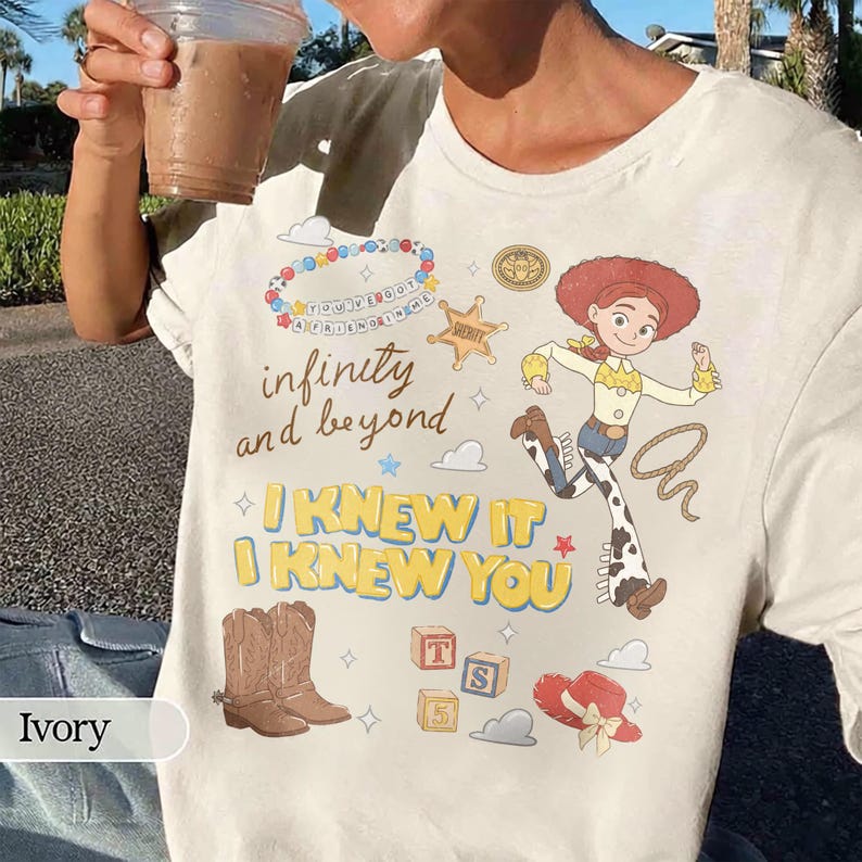 May include: An ivory t-shirt featuring a graphic print of a cowgirl character, a sheriff badge, cowboy boots, alphabet blocks, a hat, and a beaded bracelet. The text reads "infinity and beyond", "YOU'VE GOT A FRIEND IN ME", and "I KNEW IT I KNEW YOU".