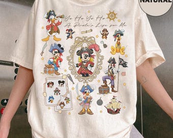 May include: Cream-colored t-shirt featuring a pirate-themed design with various cartoon characters. The design includes Mickey Mouse, Donald Duck, and Goofy dressed as pirates, along with the text "Yo Ho Yo Ho A Pirate's Life for Me."
