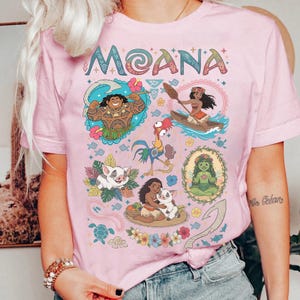 May include: Pale pink t-shirt with the word "MOANA" in a decorative font. The shirt features colourful illustrations of characters and elements from the animated film, including Maui, Moana, Hei Hei, Pua, and more. The design is surrounded by floral and tropical motifs.