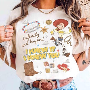 May include: A cream-colored t-shirt featuring a graphic print of Jessie the cowgirl, a sheriff badge, cowboy boots, a red hat, alphabet blocks, and a beaded bracelet. The text reads infinity and beyond, I KNEW IT, and I KNEW YOU.