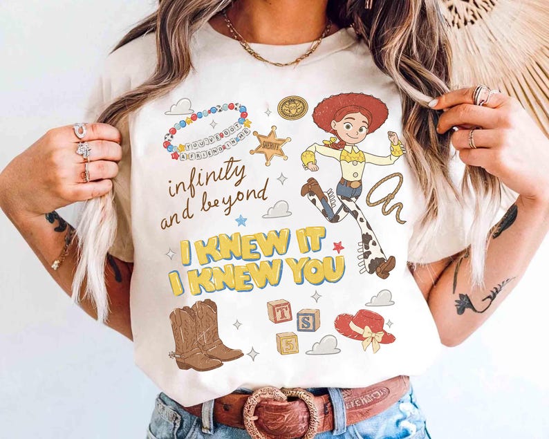May include: A cream-colored t-shirt featuring a graphic print of Jessie the cowgirl, a sheriff badge, cowboy boots, a red hat, alphabet blocks, and a beaded bracelet. The text reads infinity and beyond, I KNEW IT, and I KNEW YOU.