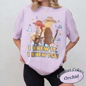 May include: A light purple short-sleeve t-shirt featuring a graphic of two figures in cowboy hats playing guitars. The text reads I KNEW IT, I KNEW YOU. The design includes small clouds, musical notes, and stars in yellow, blue, and brown tones.