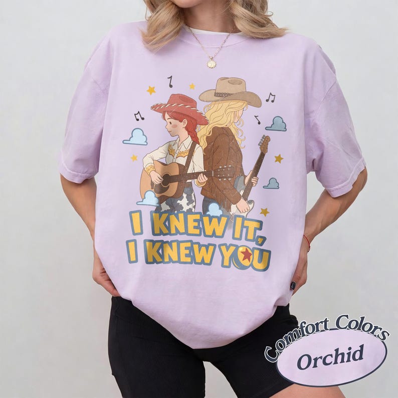 May include: A light purple short-sleeve t-shirt featuring a graphic of two figures in cowboy hats playing guitars. The text reads I KNEW IT, I KNEW YOU. The design includes small clouds, musical notes, and stars in yellow, blue, and brown tones.