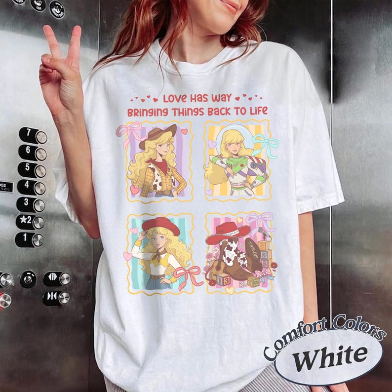 May include: A white short-sleeve t-shirt featuring a graphic print with the text LOVE HAS WAY BRINGING THINGS BACK TO LIFE. The design includes four square panels depicting a blonde character in various outfits, including a cowgirl and space ranger.