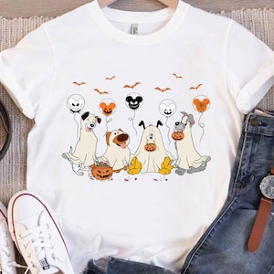 Puede incluir: Camiseta blanca con un diseño de Halloween. El diseño presenta perros de dibujos animados disfrazados de fantasmas, sosteniendo cubos y globos con forma de calabaza. Murciélagos naranjas vuelan sobre los perros. Es una prenda informal.