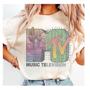 Könnte beinhalten: Cremefarbenes T-Shirt mit einem Grafikdesign im Retro-Stil des MTV-Logos und einem Kaktus-Design. Die Grafik zeigt eine Wüstenlandschaft mit Kakteen und den Worten "MUSIC TELEVISION" in einer Vintage-Schriftart.