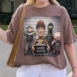 Puede incluir: Una camiseta marr&oacute;n con un estampado gr&aacute;fico de cuatro personajes de juguete en una rueda de reconocimiento policial. Buzz, Woody, Rex y Hamm sostienen carteles con sus nombres y n&uacute;meros. El fondo muestra una pared con una escala.