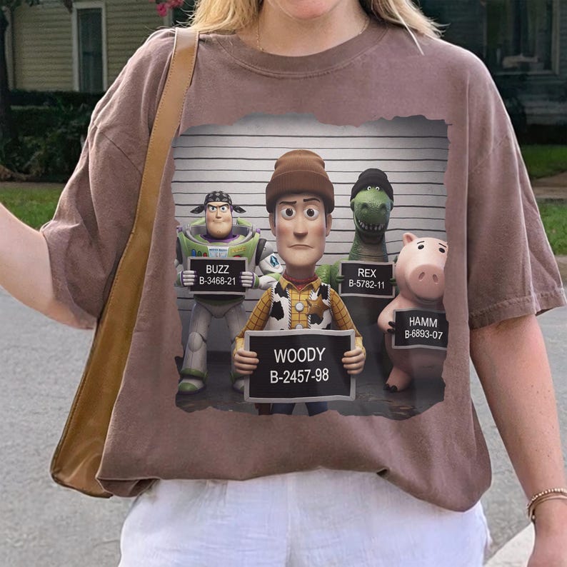 Puede incluir: Una camiseta marr&oacute;n con un estampado gr&aacute;fico de cuatro personajes de juguete en una rueda de reconocimiento policial. Buzz, Woody, Rex y Hamm sostienen carteles con sus nombres y n&uacute;meros. El fondo muestra una pared con una escala.