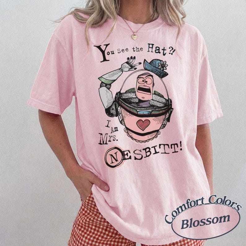 Puede incluir: Camiseta rosa claro con un dise&ntilde;o de la Sra. Nesbitt de Toy Story. El dise&ntilde;o incluye el texto "You See the Hat? I Am Mrs. Nesbitt!" y un coraz&oacute;n. La camiseta est&aacute; etiquetada como "Comfort Colors Blossom".