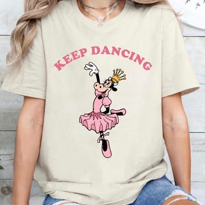 May include: Cream-colored t-shirt featuring a cartoon cow in a pink ballerina outfit, wearing a gold crown, and dancing. The words "KEEP DANCING" are printed above the cow in pink letters. The shirt has short sleeves.