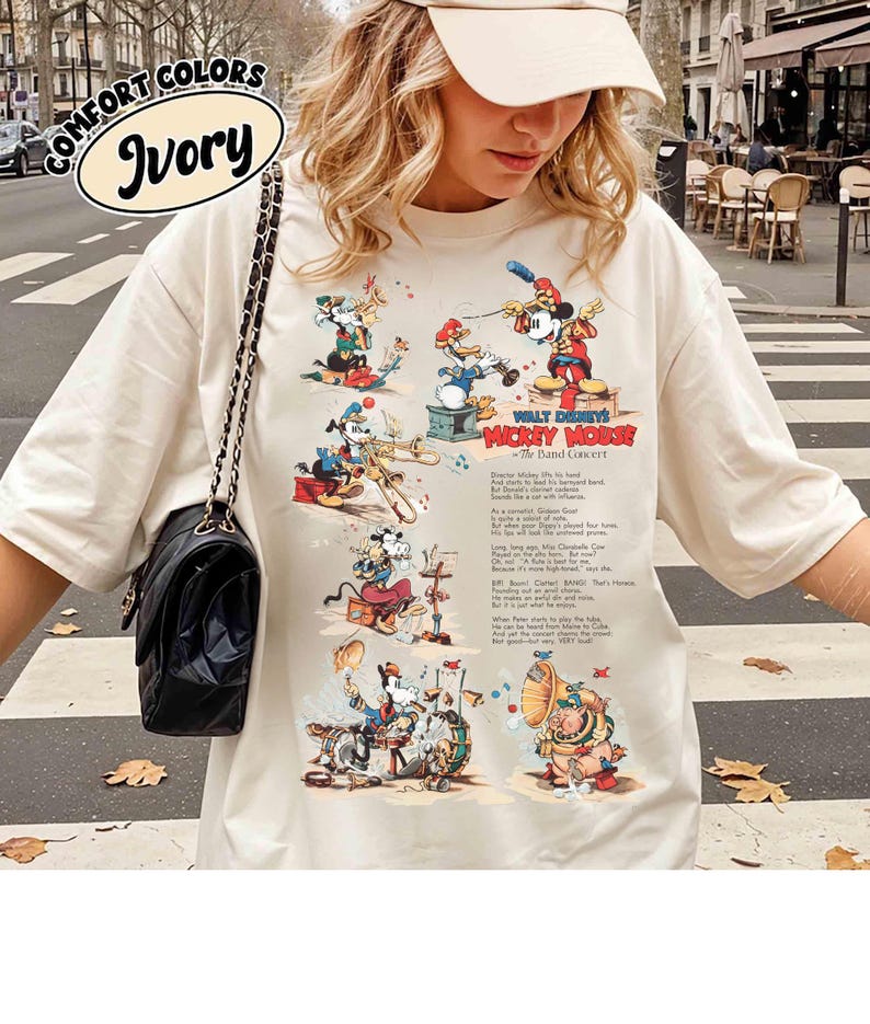 Puede incluir: Camiseta de color marfil con una escena de banda de dibujos animados vintage con Mickey Mouse y sus amigos. La camiseta muestra el texto "WALT DISNEY'S MICKEY MOUSE - The Band Concert" y "COMFORT COLORS Ivory" en un &oacute;valo. El dise&ntilde;o es en rojo, azul y amarillo.