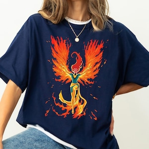 Può includere: T-shirt blu navy con una grafica di una fenice con ali arancioni e testa rossa. La figura della fenice ha le braccia tese e un design giallo e oro. La maglietta ha un colletto bianco.