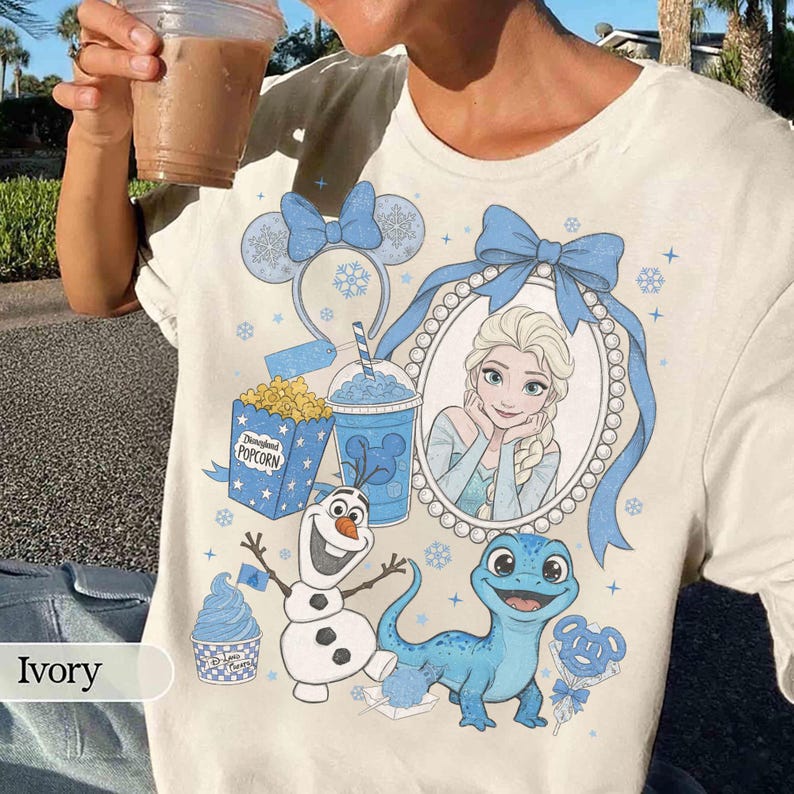 Op de afbeelding: Ivoor kleurig T-shirt met een Disney-thema ontwerp. De afbeelding bevat Elsa, Olaf de sneeuwman en een blauwe hagedis. Extra elementen zijn popcorn, een slushy en een hoofdband met muisoren.