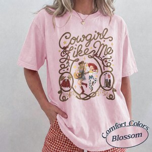 May include: A light pink t-shirt featuring the text Cowgirl like Me in brown script. The graphic includes illustrations of two female characters, a guitar, a cowboy hat, a jacket, and boots, all framed by a rope design with musical notes.