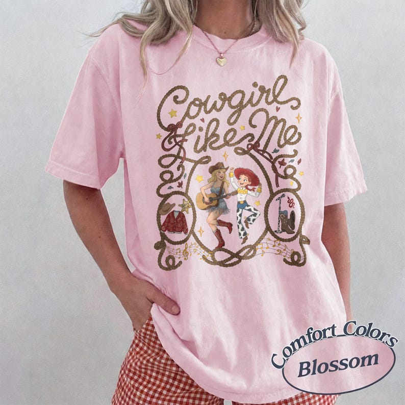May include: A light pink t-shirt featuring the text Cowgirl like Me in brown script. The graphic includes illustrations of two female characters, a guitar, a cowboy hat, a jacket, and boots, all framed by a rope design with musical notes.