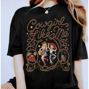 May include: A black t-shirt featuring the text Cowgirl Like Me in a rope-style font. The graphic includes two female characters, one playing an acoustic guitar, alongside illustrations of a cowboy hat, a jacket, a sheriff badge, and a boot.