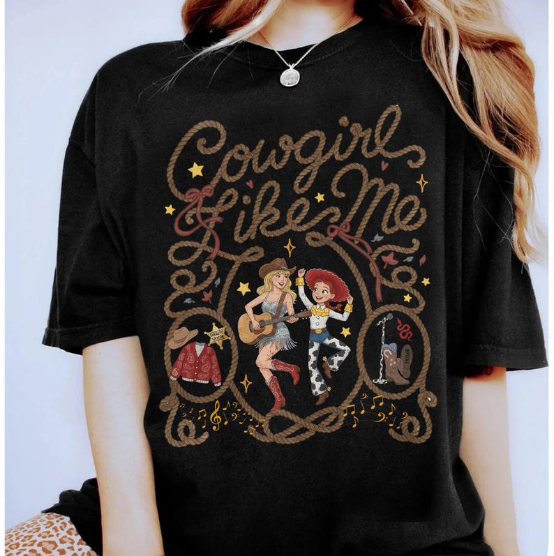 May include: A black t-shirt featuring the text Cowgirl Like Me in a rope-style font. The graphic includes two female characters, one playing an acoustic guitar, alongside illustrations of a cowboy hat, a jacket, a sheriff badge, and a boot.