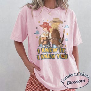 May include: A light pink short-sleeve t-shirt featuring a graphic illustration of two figures wearing cowboy hats and playing acoustic guitars. The text "I KNEW IT, I KNEW YOU" is printed in yellow and blue below the figures, surrounded by stars and clouds.
