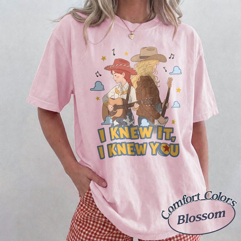 May include: A light pink short-sleeve t-shirt featuring a graphic illustration of two figures wearing cowboy hats and playing acoustic guitars. The text "I KNEW IT, I KNEW YOU" is printed in yellow and blue below the figures, surrounded by stars and clouds.