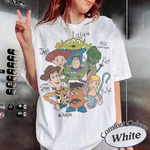 May include: White t-shirt featuring a colorful graphic of characters from the animated movie Toy Story. The design includes Woody, Buzz Lightyear, Jessie, Rex, Mr. Potato Head, Bo Peep, and an Alien. The shirt is labeled "Comfort Colors White".