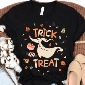May include: Black t-shirt with a Halloween graphic. The design includes the words "Trick or Treat" in orange and white, alongside a cartoon ghost carrying a candy basket. Autumn leaves and stars complete the festive design.